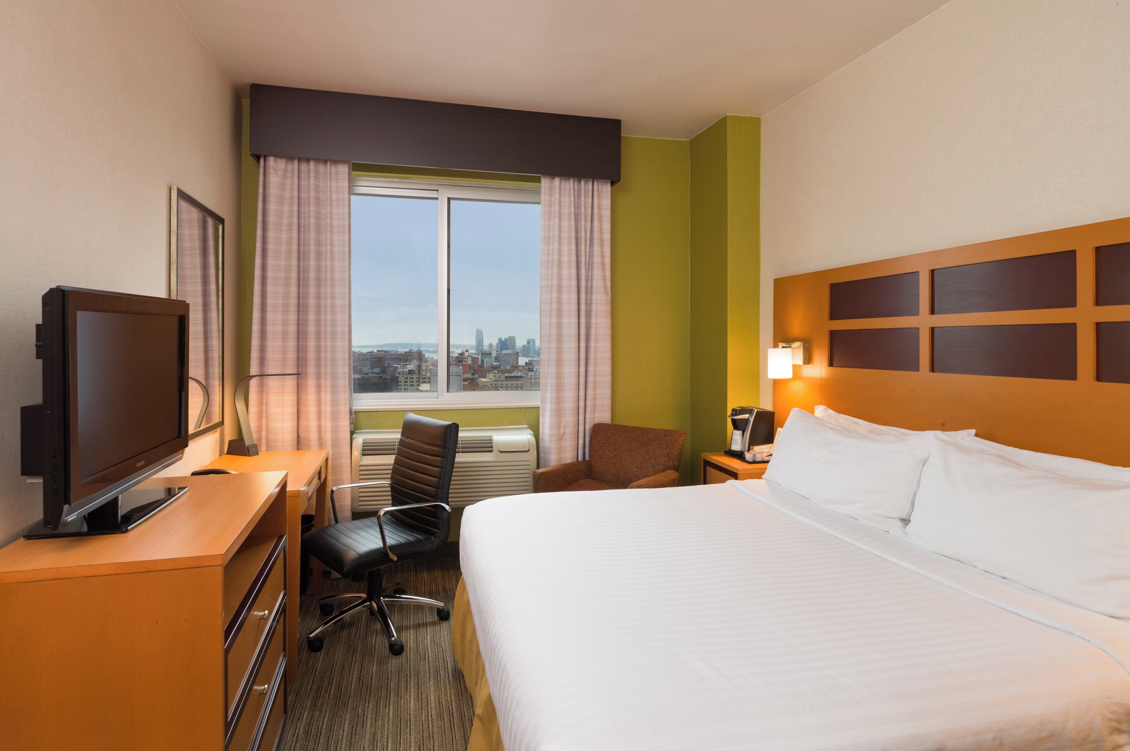 A hotel room with a bed, a desk with a television, a chair, a window with curtains, and a city view.