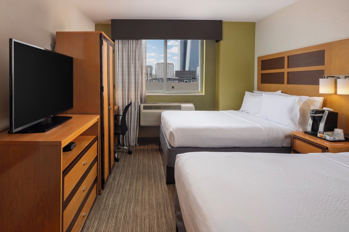 A hotel room with two beds, a TV on a wooden dresser, a window with a city view, and a bedside table with a lamp and a coffee maker.