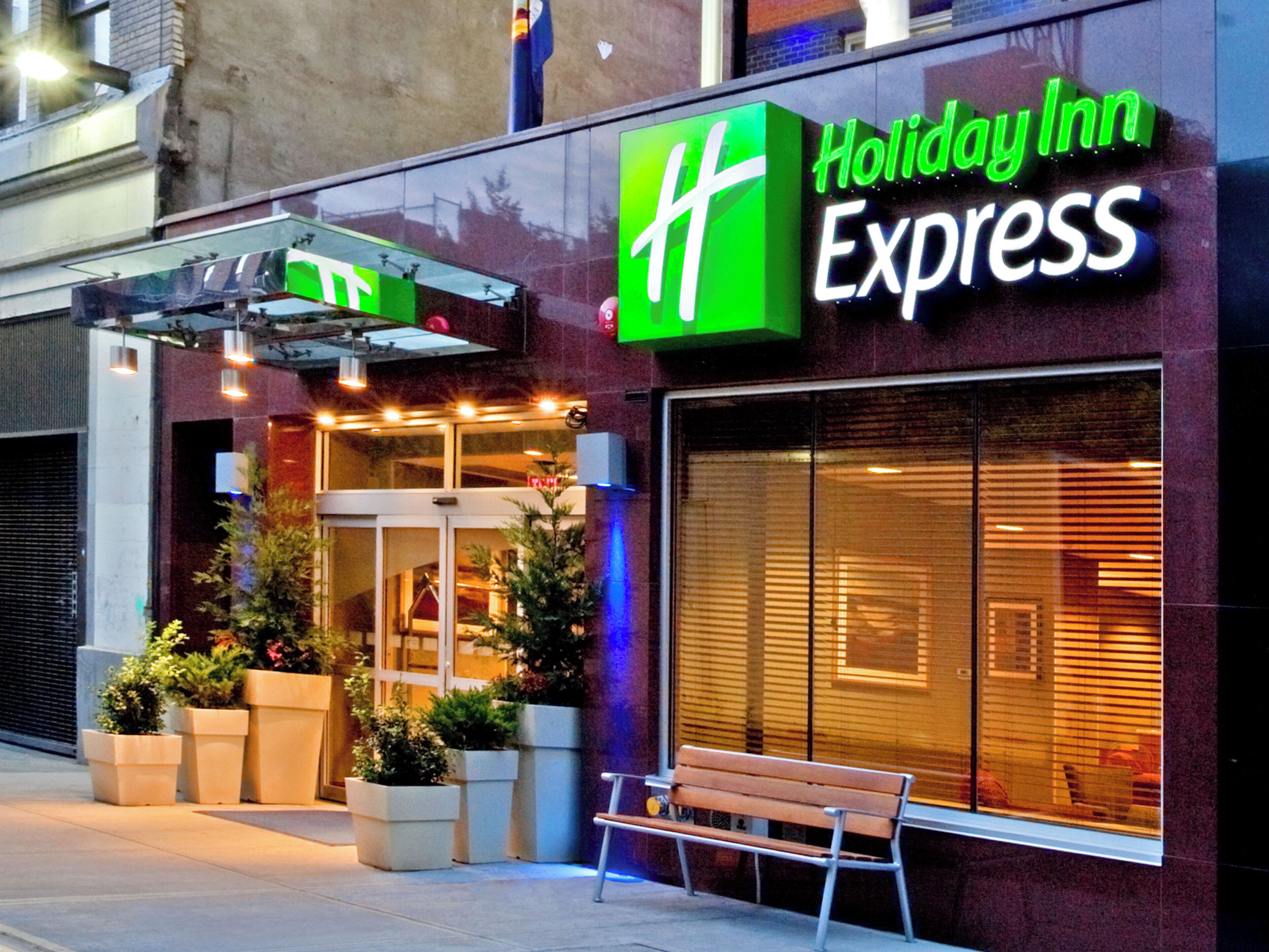 Un hôtel Holiday Inn Express reconnaissable à son logo vert bien visible, à son entrée en verre et aux plantes en pot qui l’ornent à l’extérieur.