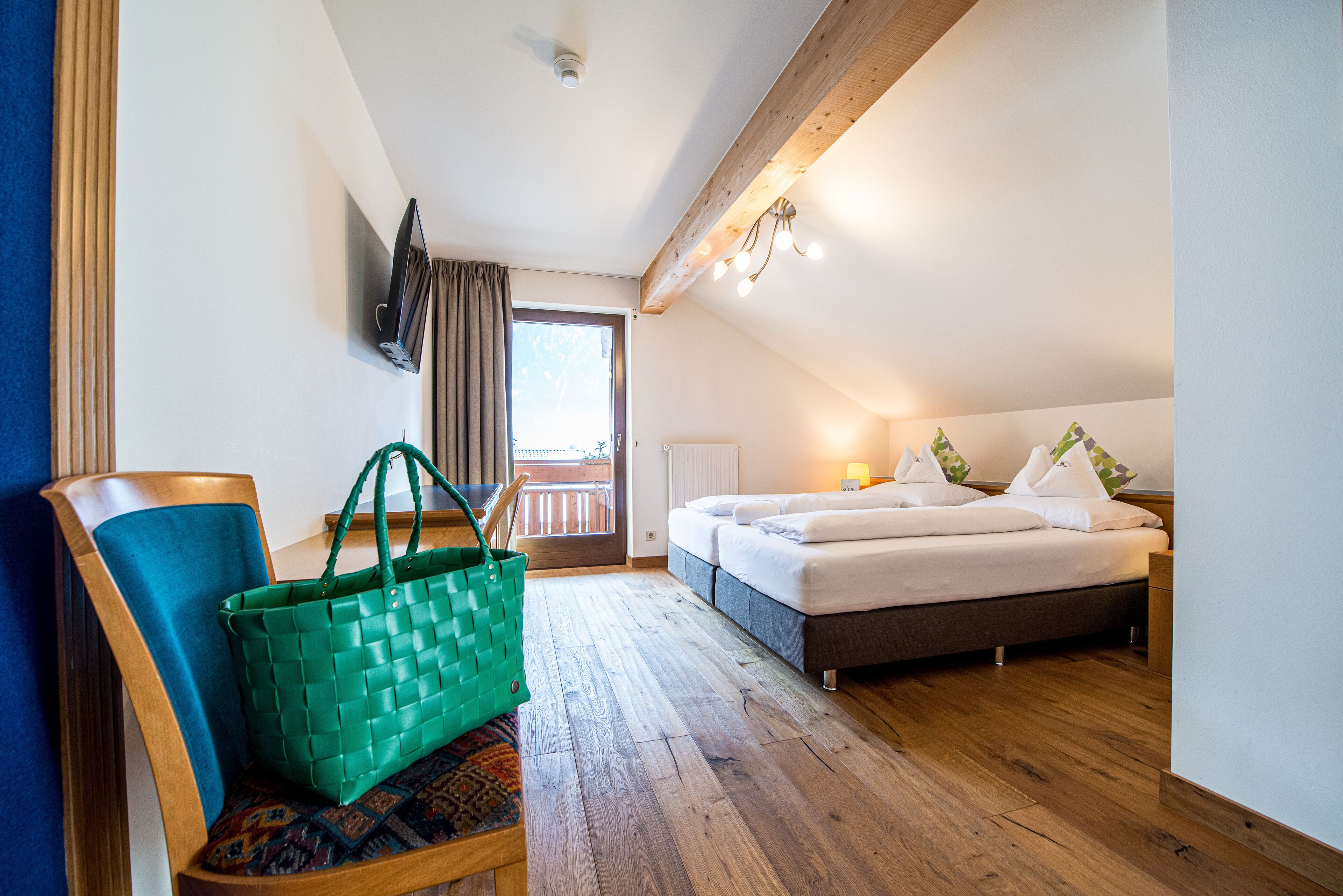 Una habitación de hotel con piso de madera, una cama con sábanas blancas, un bolso verde sobre una silla y un televisor montado en la pared.