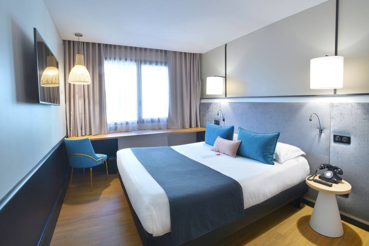 A modern hotel room with a large bed, a desk, a chair, and a bedside table with a telephone.