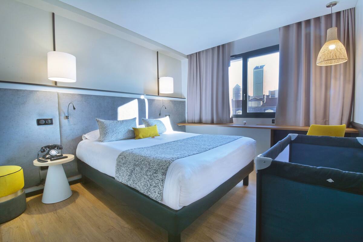 A modern hotel room with a large bed, bedside tables, a desk, and a view of the city.