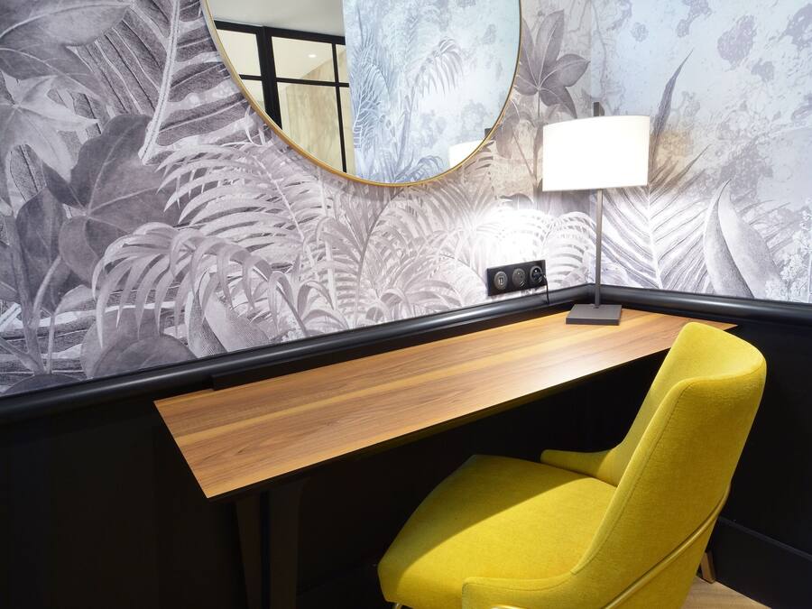 Una habitación con escritorio, una silla amarilla, un espejo redondo y una pared con diseño de hojas.