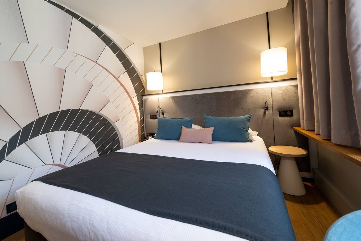 A modern hotel room with a large bed, a round wall clock, a bedside table, and a small round table.