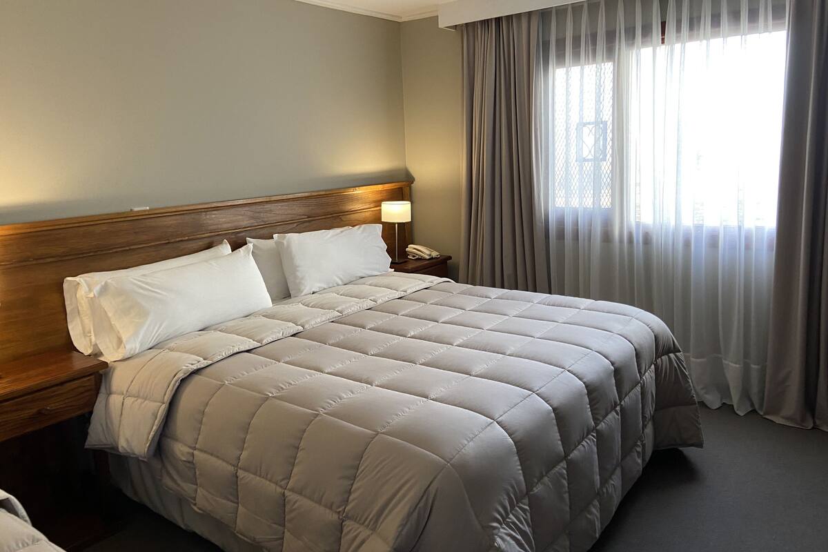 Hotelkamer met een groot bed, houten hoofdbord, nachtkastje met lamp en raam met dunne gordijnen.