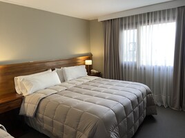 Habitación de hotel con una cama grande, cabecera de madera, una mesita de noche con lámpara y una ventana con cortinas traslúcidas.