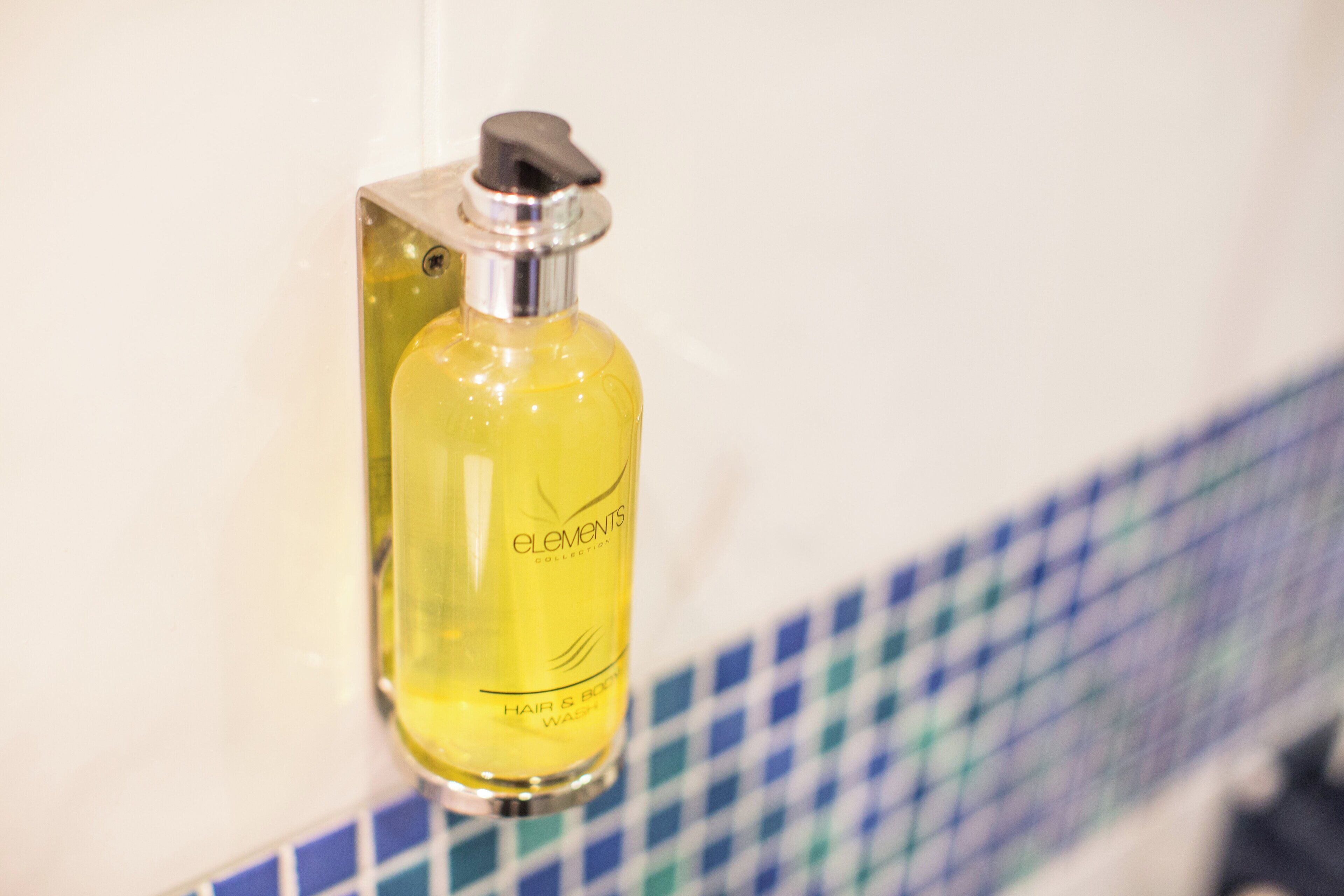 A yellow bottle of Elements Collection hair and body wash mounted on a wall with a blue and white checkered tile backsplash.