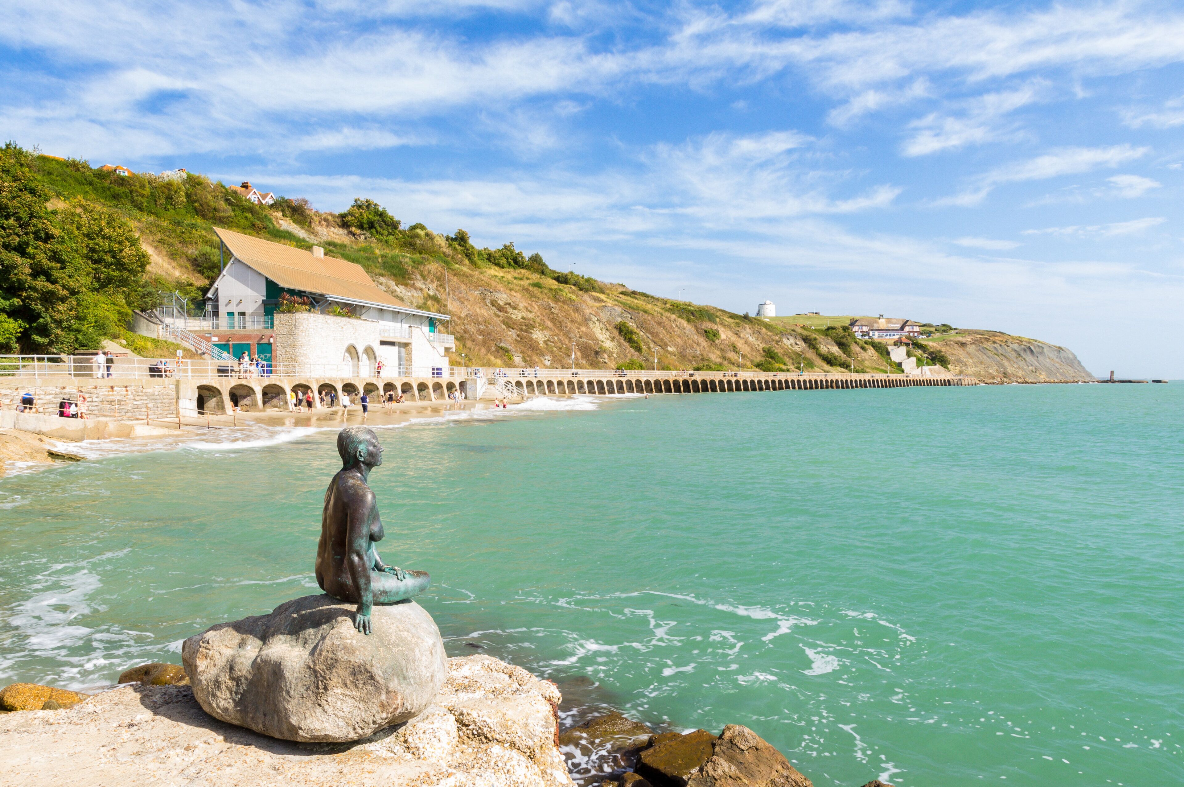 A coastal scene with a statue on a rock, a building with a sloped roof, and a pier extending into the water.