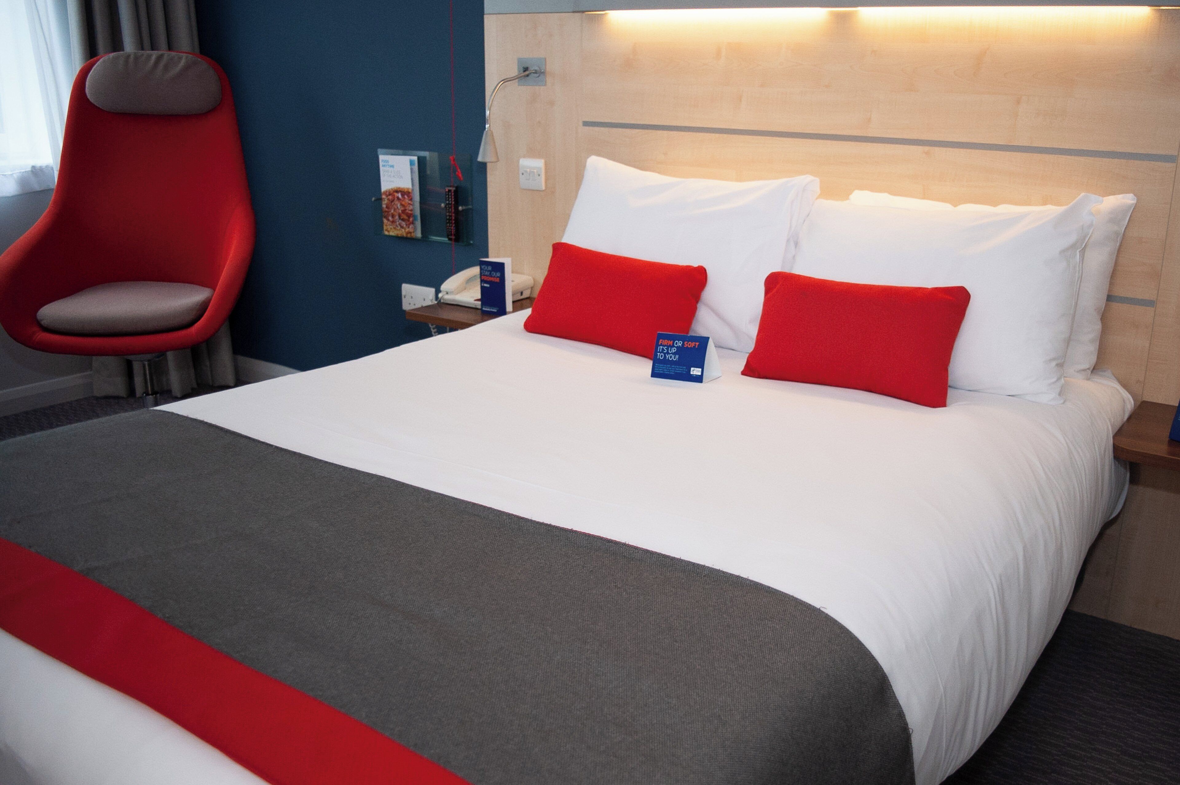 A hotel room with a bed, a red chair, a bedside table with a phone, and a sign indicating room firmness.