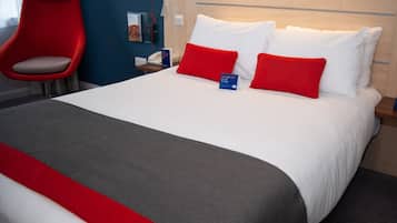 Habitación de hotel con una cama grande, almohadas rojas y blancas, una silla roja, una mesita de noche con una lámpara y un cartel en la cama.
