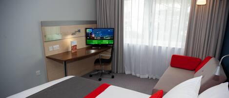 A hotel room with a bed, a desk with a computer, a chair, and a window with curtains.