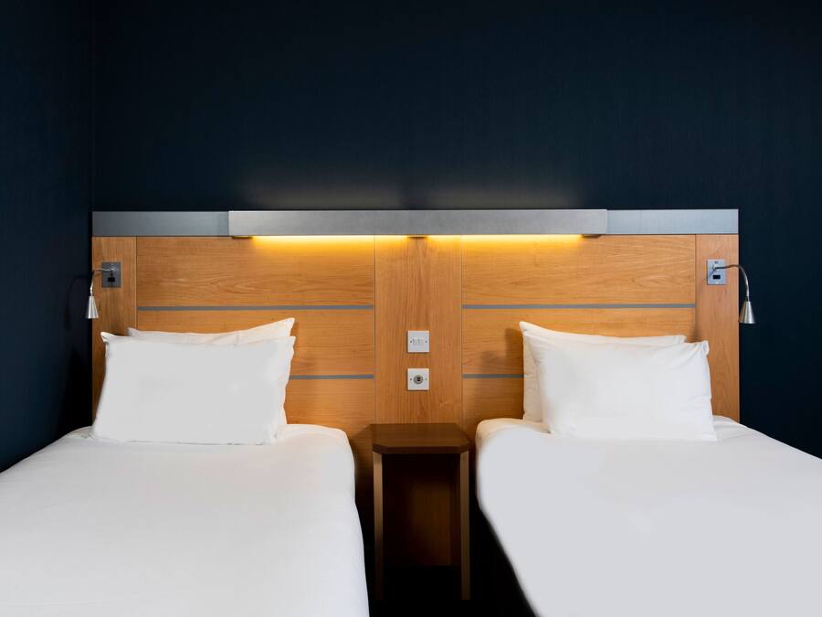 Ein Hotelzimmer mit zwei Einzelbetten, einem hölzernen Kopfstück, Nachttischlampen und einem kleinen Nachttisch.