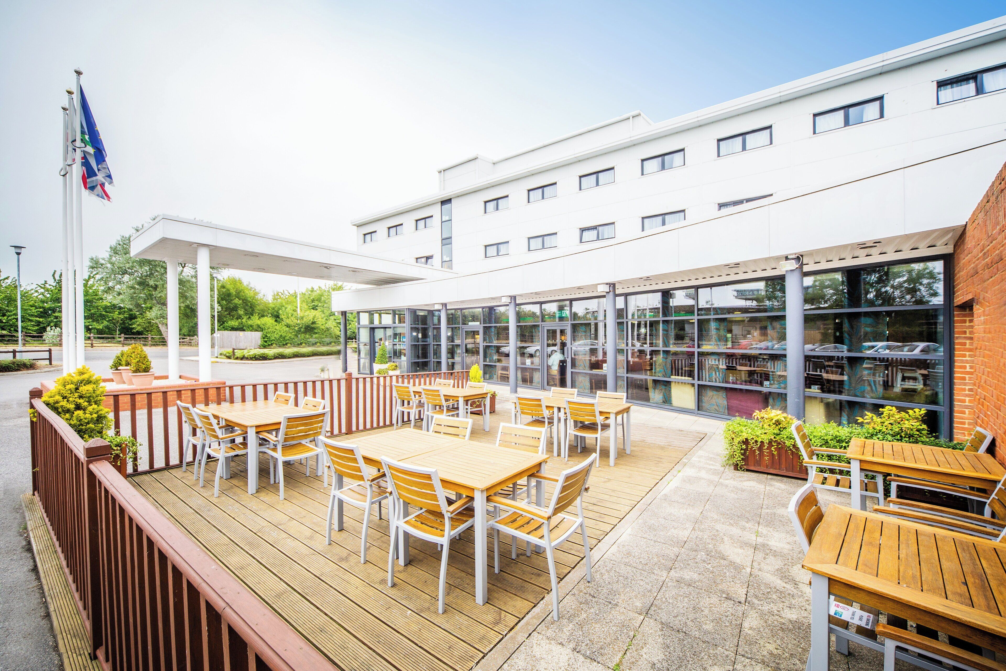 An outdoor patio area with wooden tables and chairs, a covered seating area, and a building with large windows in the background.