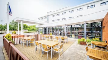 Un espace extérieur aménagé en terrasse, avec des tables et des chaises en bois, un coin salon abrité et, au fond, un bâtiment doté de grandes fenêtres.