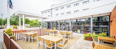 An outdoor patio area with wooden tables and chairs, a covered seating area, and a building with large windows in the background.