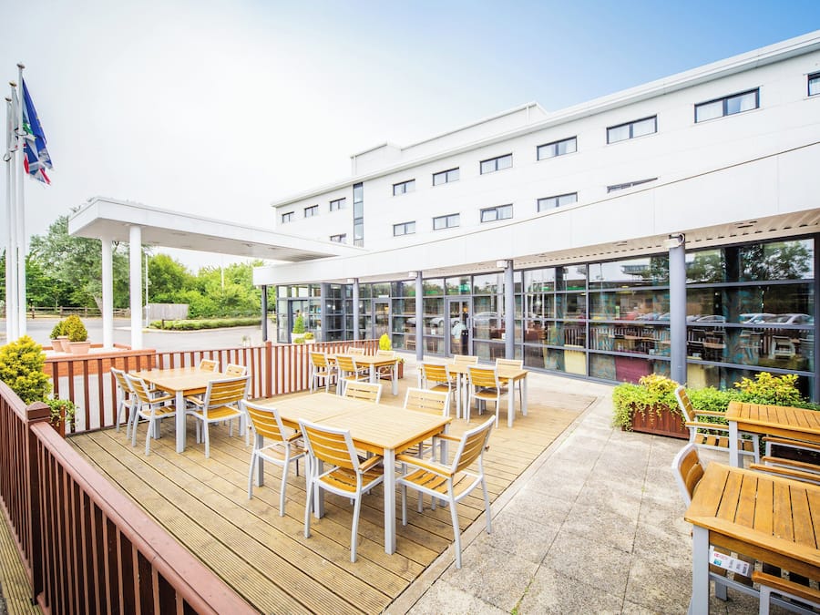An outdoor patio area with wooden tables and chairs, a covered seating area, and a building with large windows in the background.