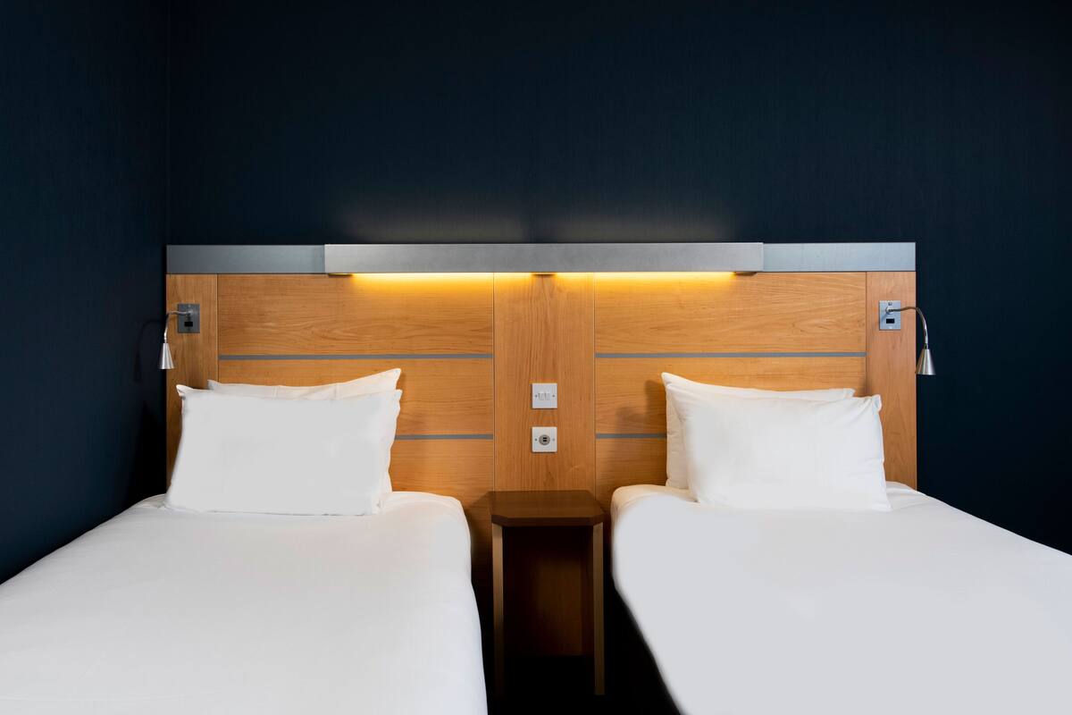 A hotel room with two single beds, wooden headboard, bedside lamps, and a small wooden nightstand.