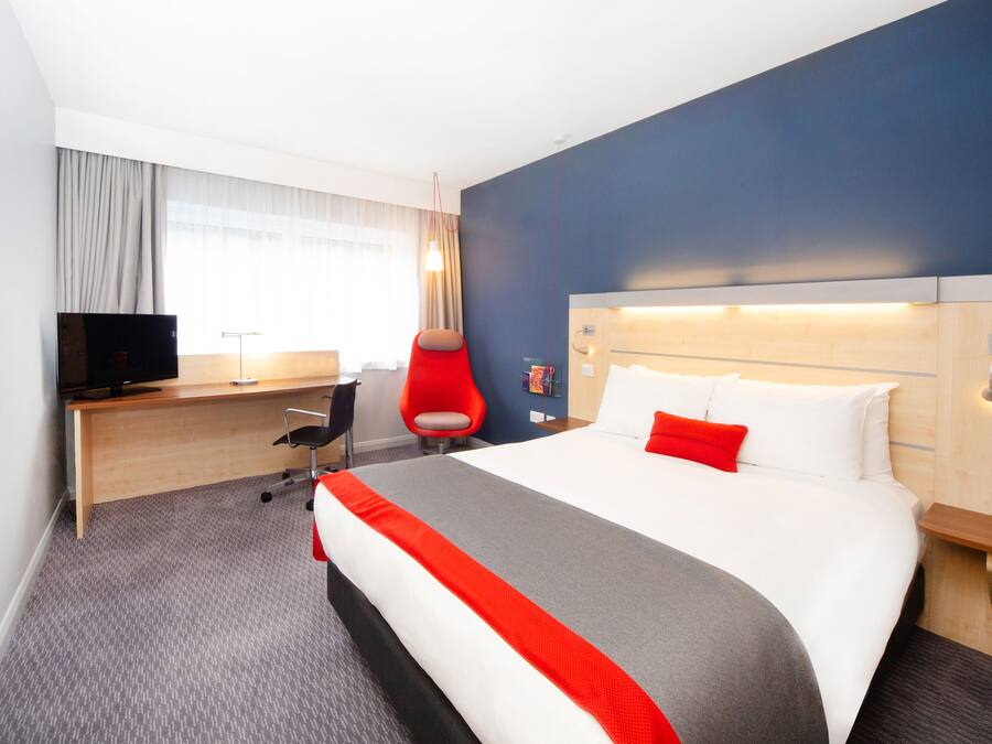 A hotel room with a large bed, a desk with a computer, a TV, and a red chair.