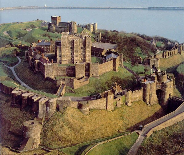 A historic castle with multiple towers and walls, surrounded by grassy hills and a body of water.