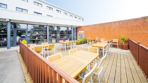 An outdoor patio with wooden tables and chairs, a brick wall, and a modern building with large windows in the background. - Holiday Inn Express Folkestone - Channel Tunnel by IHG (Folkestone)