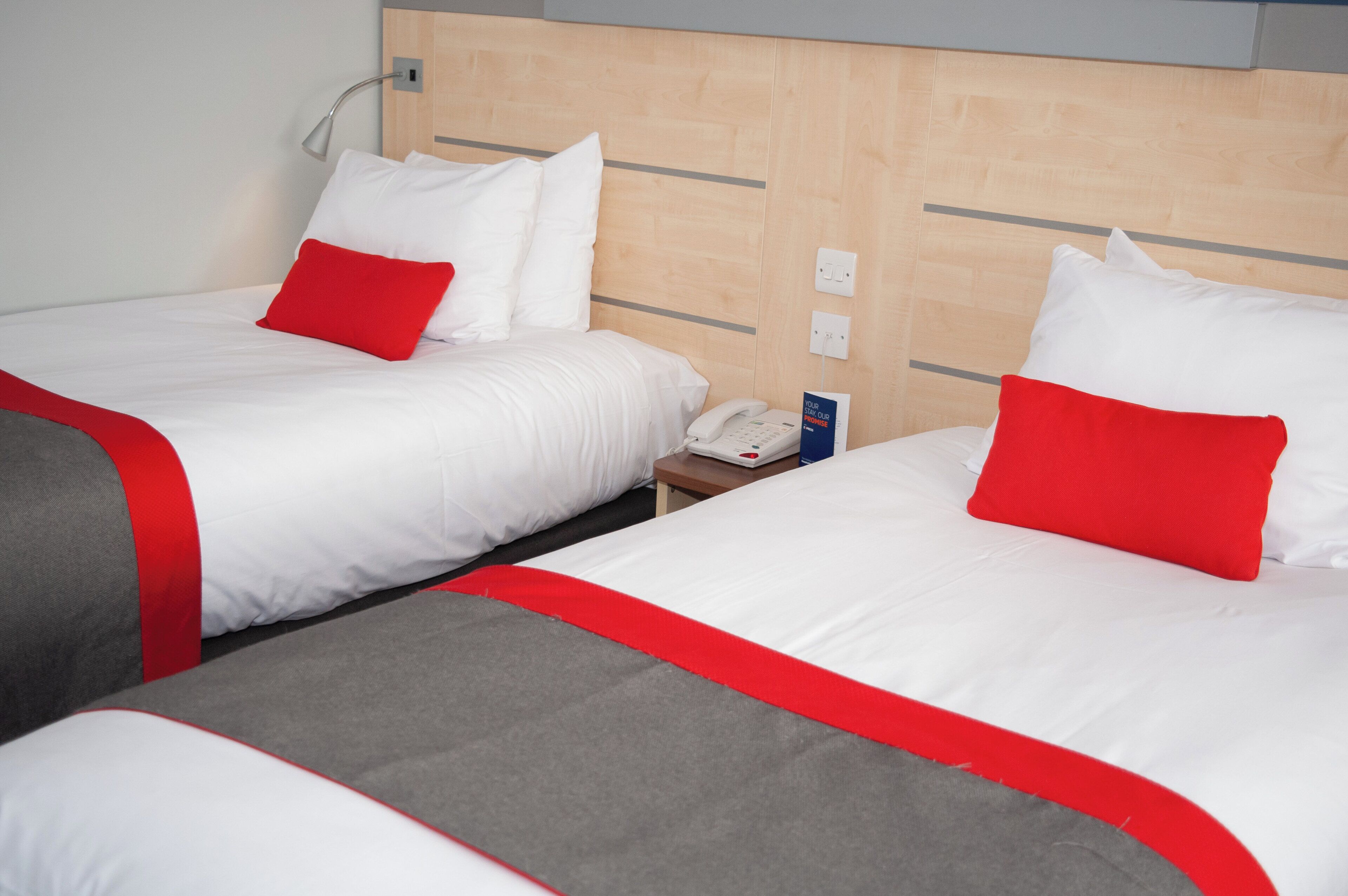 A hotel room with two beds, each with white bedding and red accents, a bedside table with a phone, and a wooden headboard.