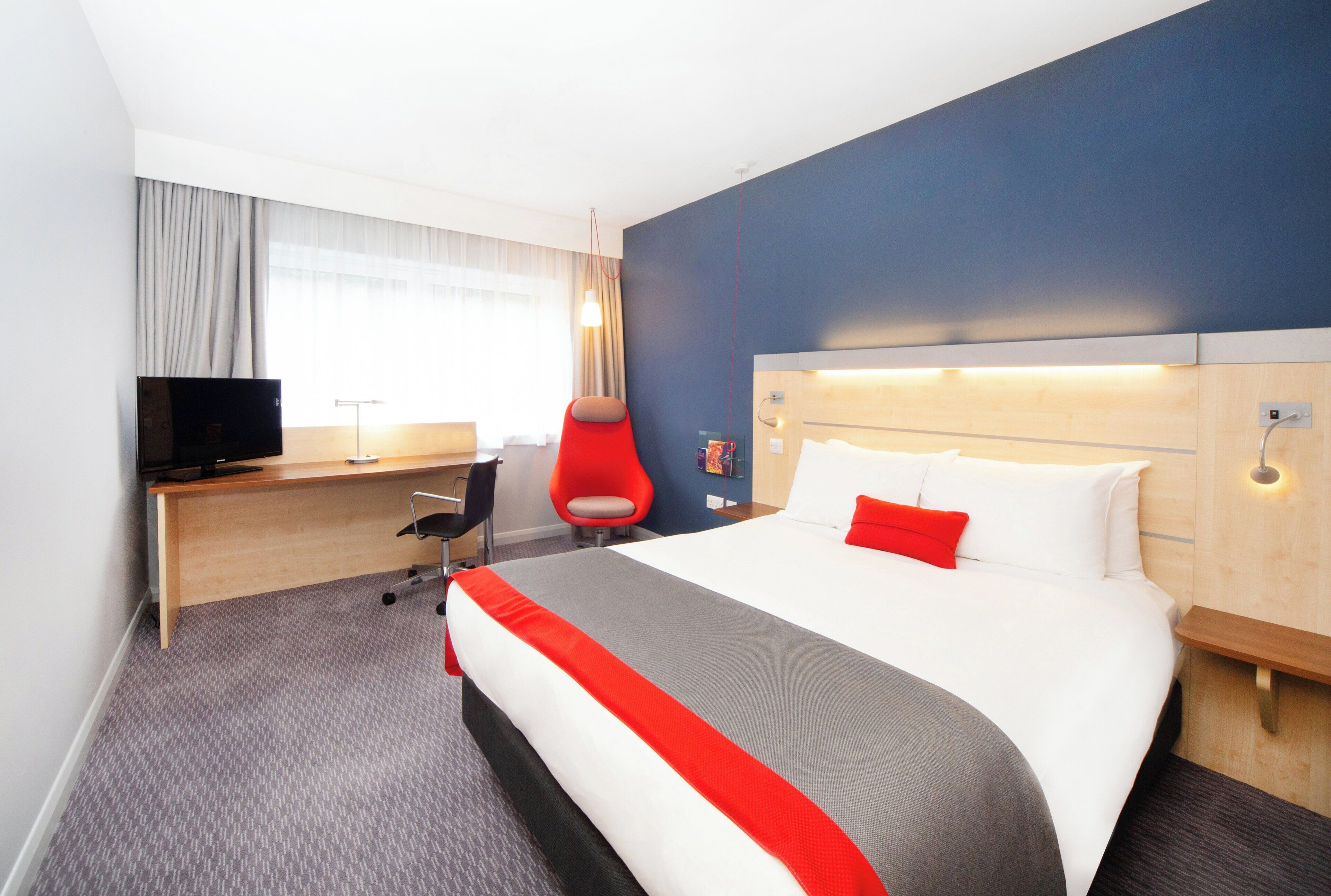 Habitación de hotel con cama, escritorio, silla, televisión y ventana con cortinas.