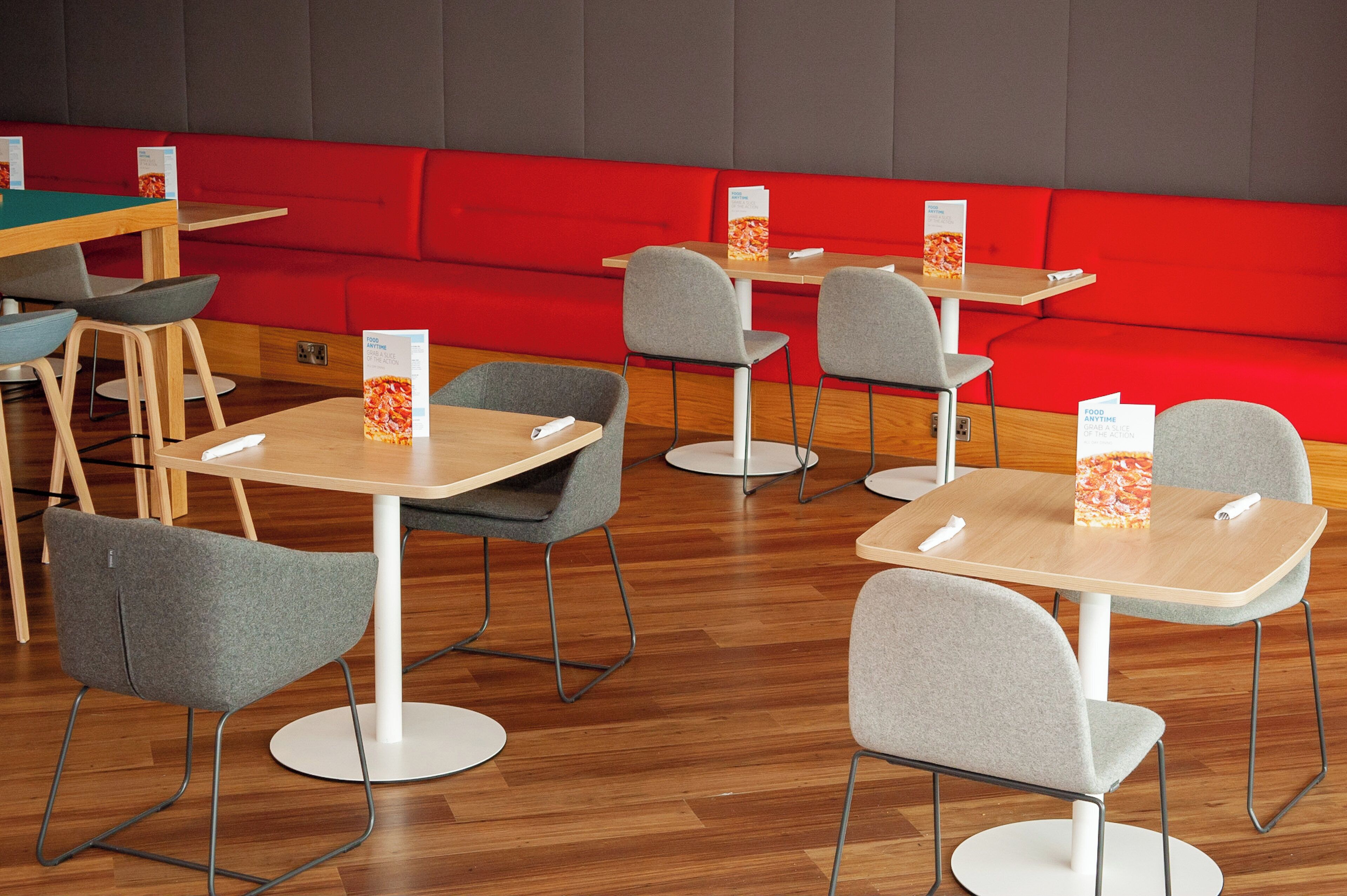 Interior de un restaurante con varias mesas y sillas, asientos con cojines rojos y atril para menús.