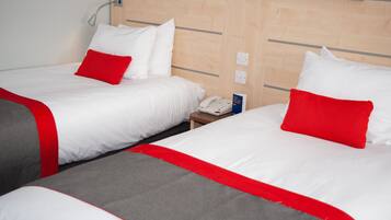 Habitación de hotel con dos camas, cada una con ropa de cama blanca y detalles en rojo. Se observa una mesita de noche con un teléfono y un cartel, además de una lámpara fijada a la pared.