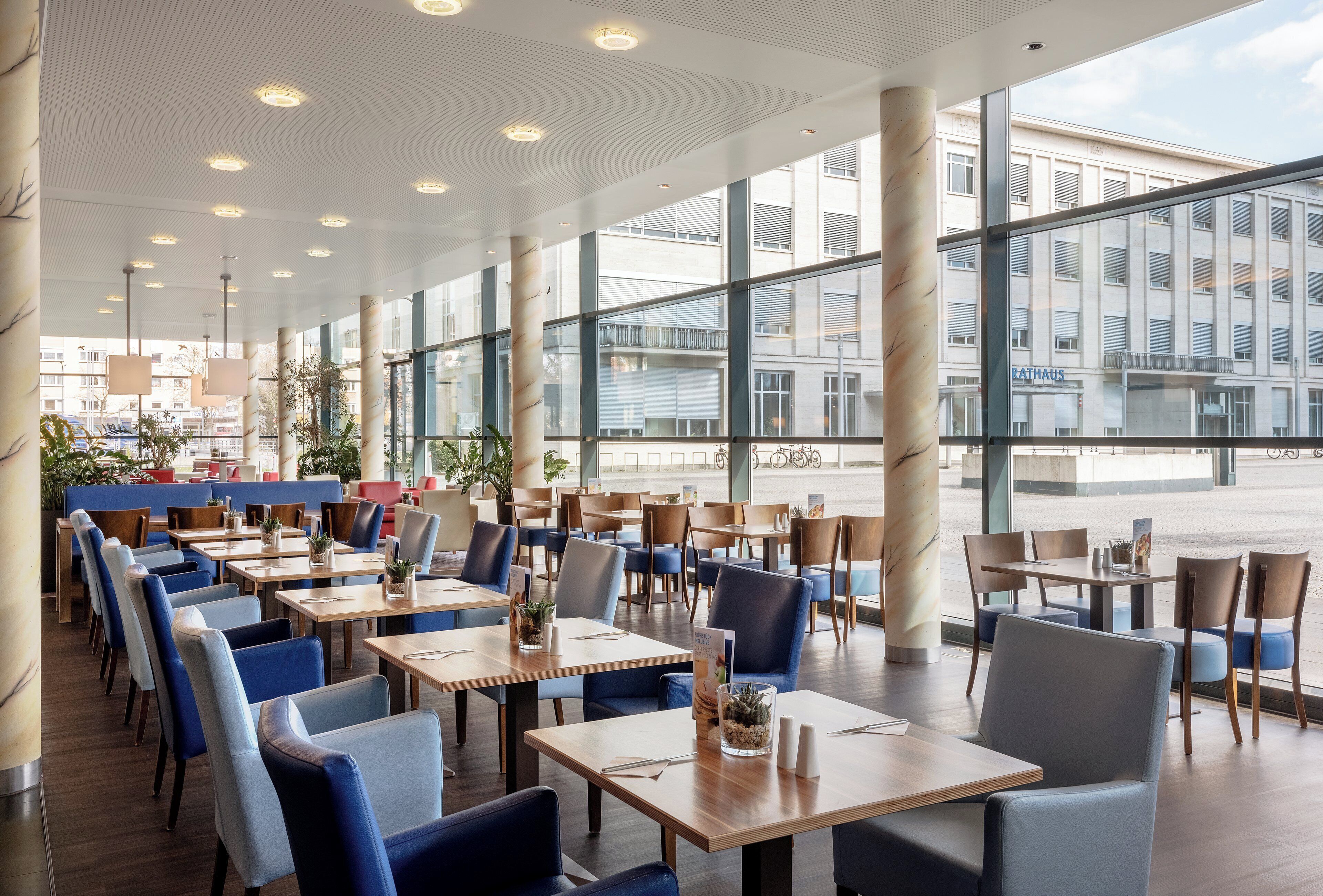 A spacious indoor dining area with multiple tables and chairs, large windows offering views of the street, and a ceiling with recessed lighting.