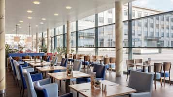 A spacious indoor dining area with multiple tables and chairs, large windows offering views of the street, and a ceiling with recessed lighting.