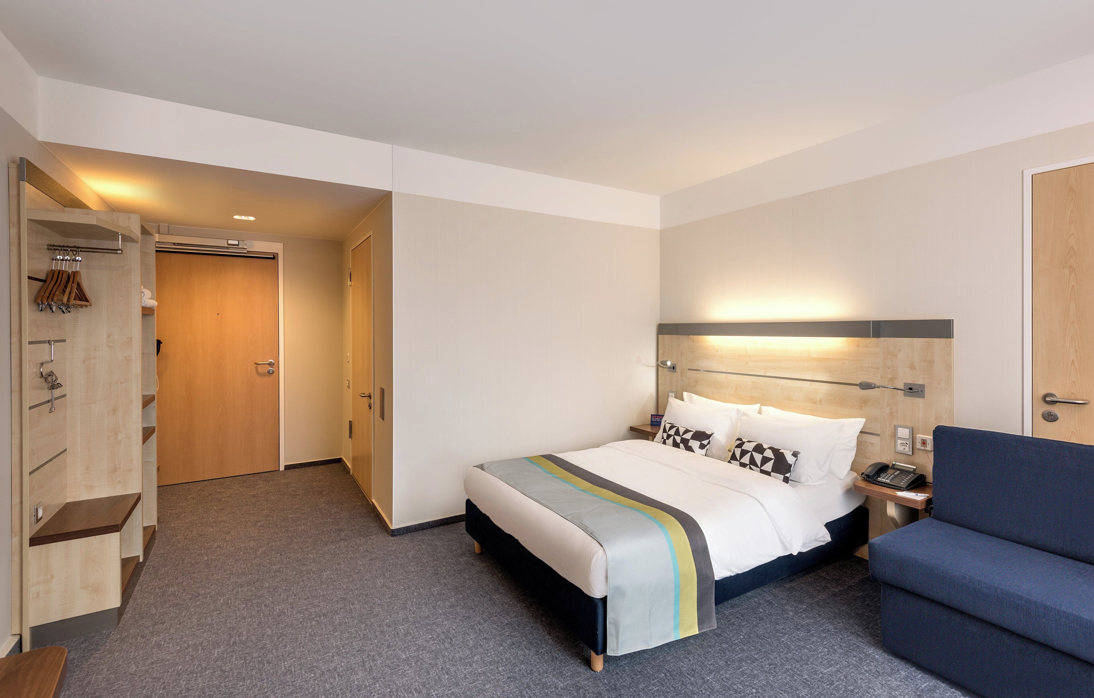 Une chambre d’hôtel comprenant un lit, une table de chevet avec un téléphone, une chaise, une armoire et une porte donnant sur une autre pièce.