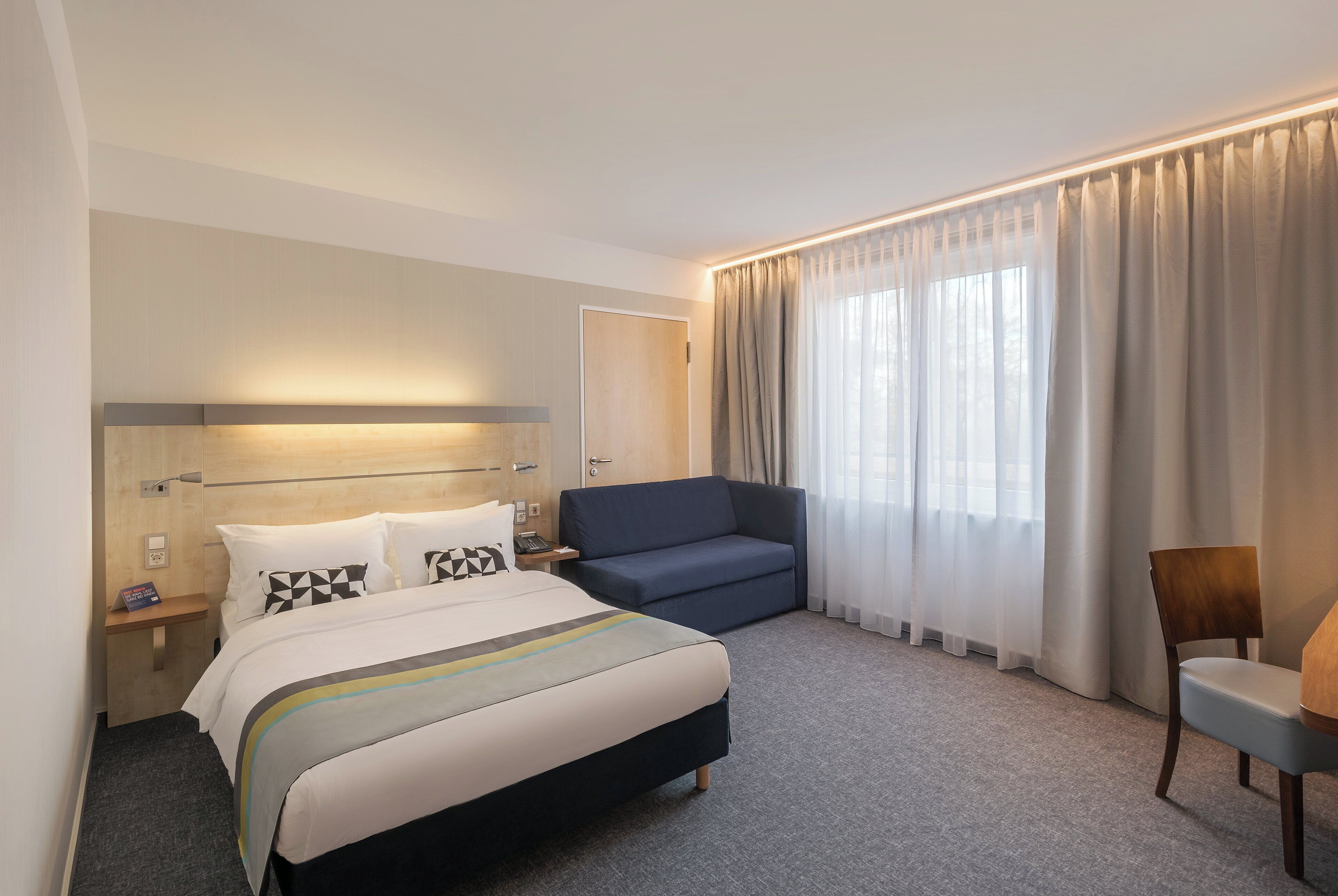 Habitación de hotel con cama, sofá, sillón, escritorio y ventana con cortinas.