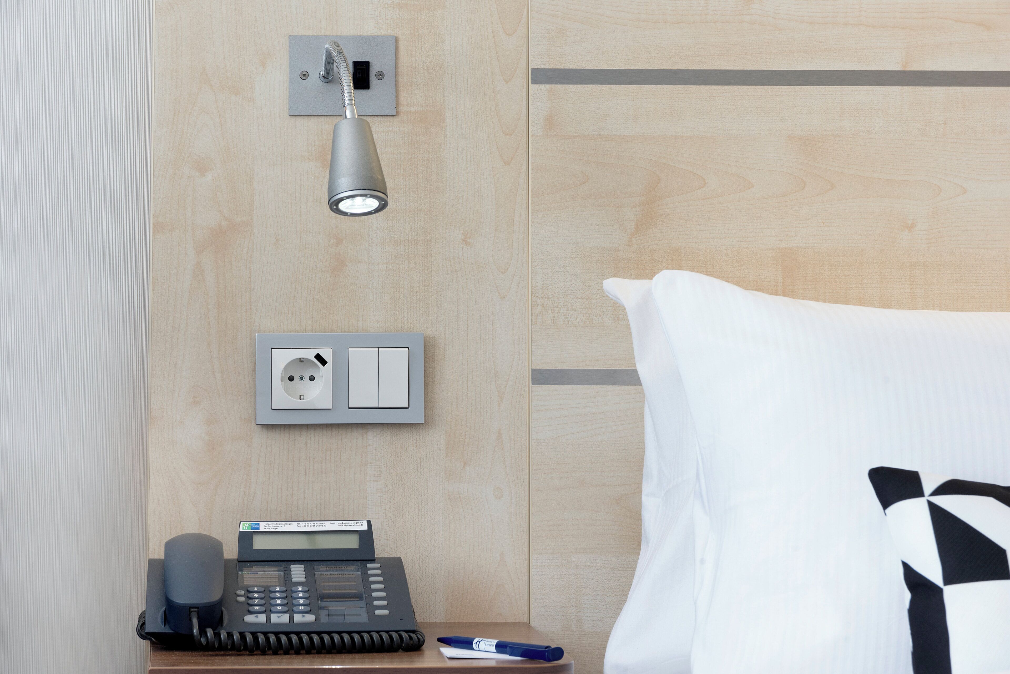Habitación de hotel con teléfono en la mesita de noche, toma de corriente e iluminación empotrada en la pared.