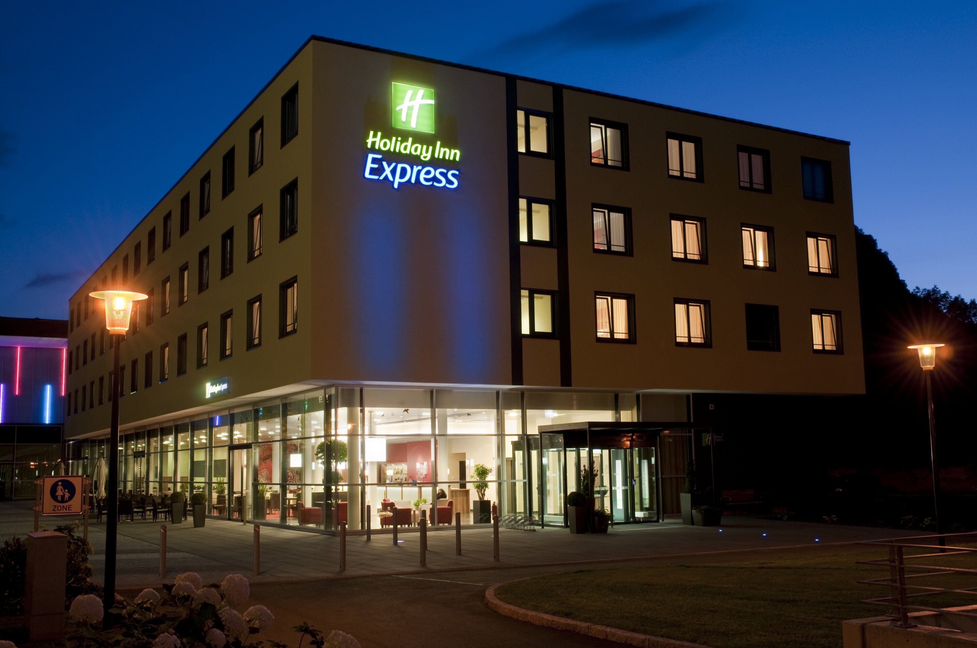 Un hotel Holiday Inn Express con fachada de vidrio, varios pisos y una entrada bien iluminada.