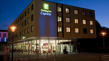 Un hotel Holiday Inn Express con fachada de vidrio, varios pisos y una entrada bien iluminada.