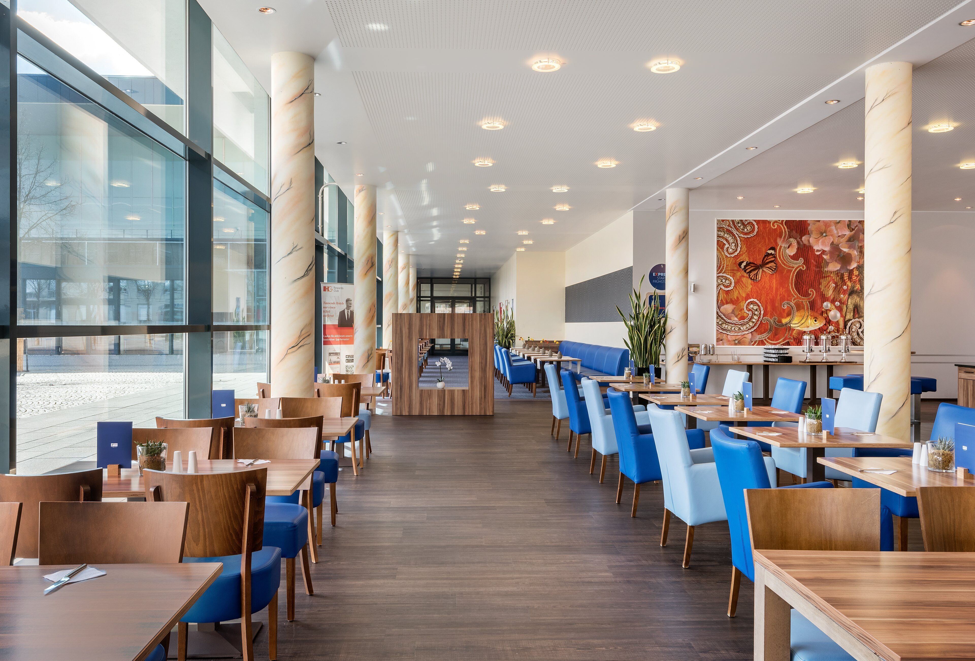 Een modern restaurant met een lange toonbank, blauwe stoelen en een groot raam.