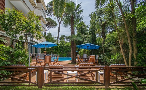 A poolside area with lounge chairs and umbrellas, surrounded by lush greenery and palm trees. - Hotel Kyrton (Forte dei Marmi)