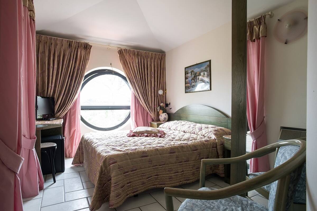 A bedroom with a bed, a desk with a chair, a window with curtains, and a wall-mounted fan.
