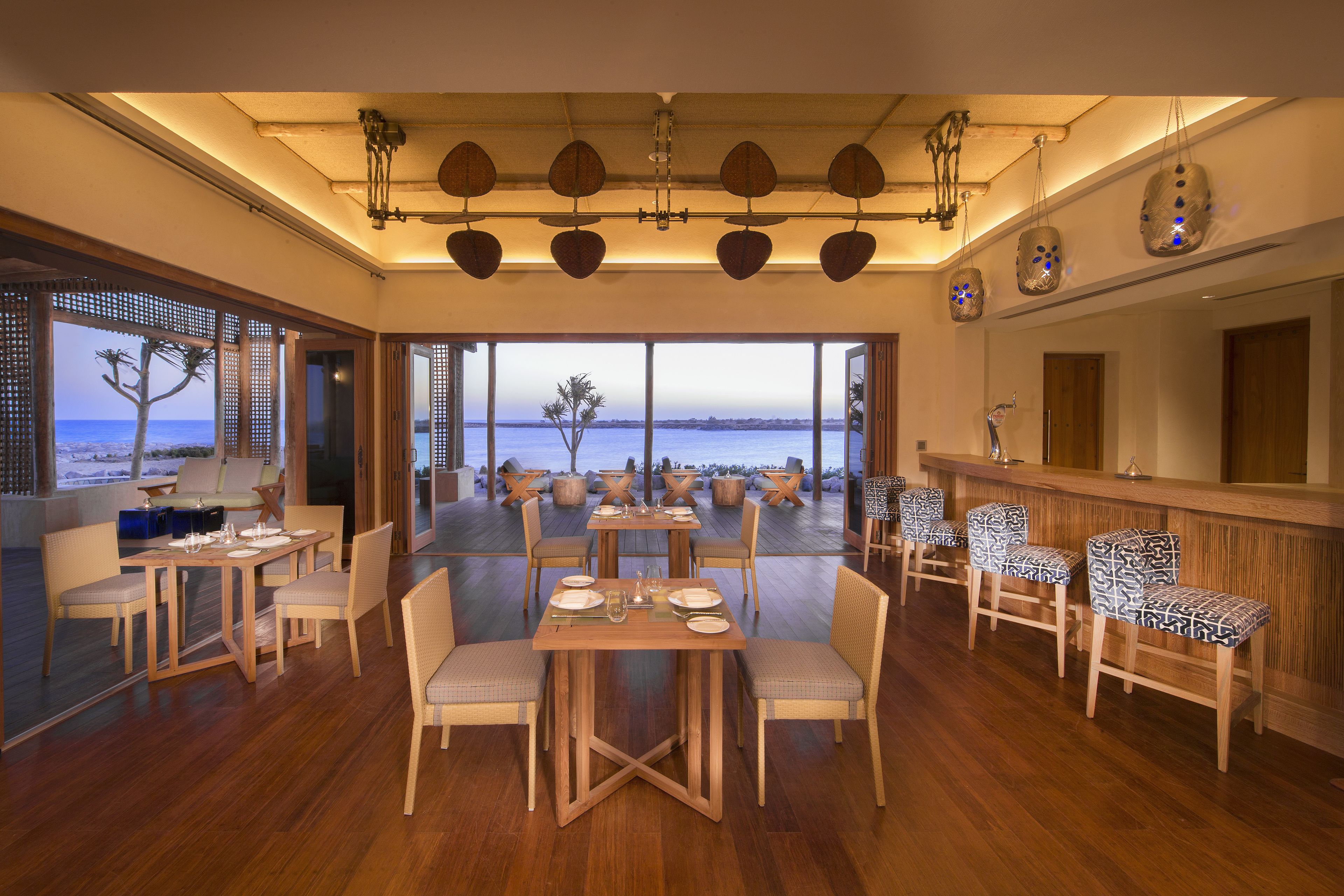 Ein geräumiger Essbereich mit Holztischen und -stühlen, große Fenster mit Blick auf den Strand und eine Bar mit gestreiften Barhocker.