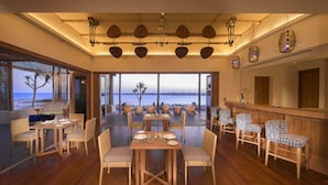 Un vaste espace dédié à la salle à manger, avec des tables et des chaises en bois, de grandes fenêtres offrant une vue sur la plage, et un coin bar agrémenté de tabourets à motifs.