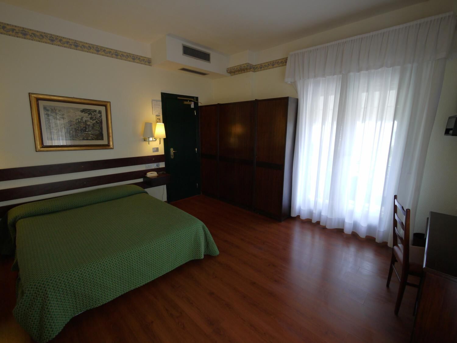Een hotelkamer met een bed, een kledingkast, een bureau met een lamp, een stoel en een raam met gordijnen.