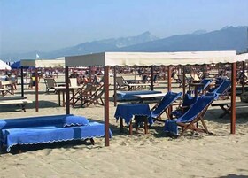 Una playa con filas de sillas de playa y mesas bajo sombrillas, y montañas al fondo.