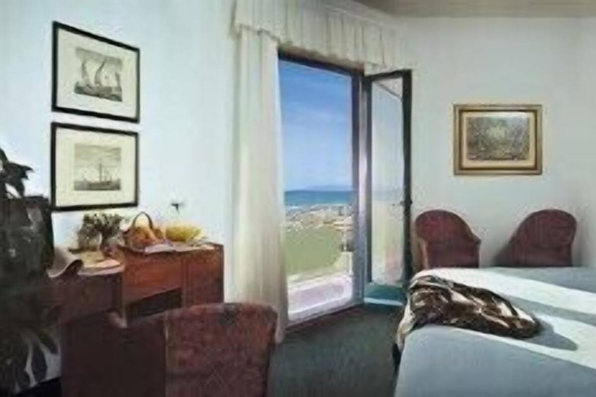 A hotel room with a bed, a desk, and a chair. There is a window with a view of the ocean.