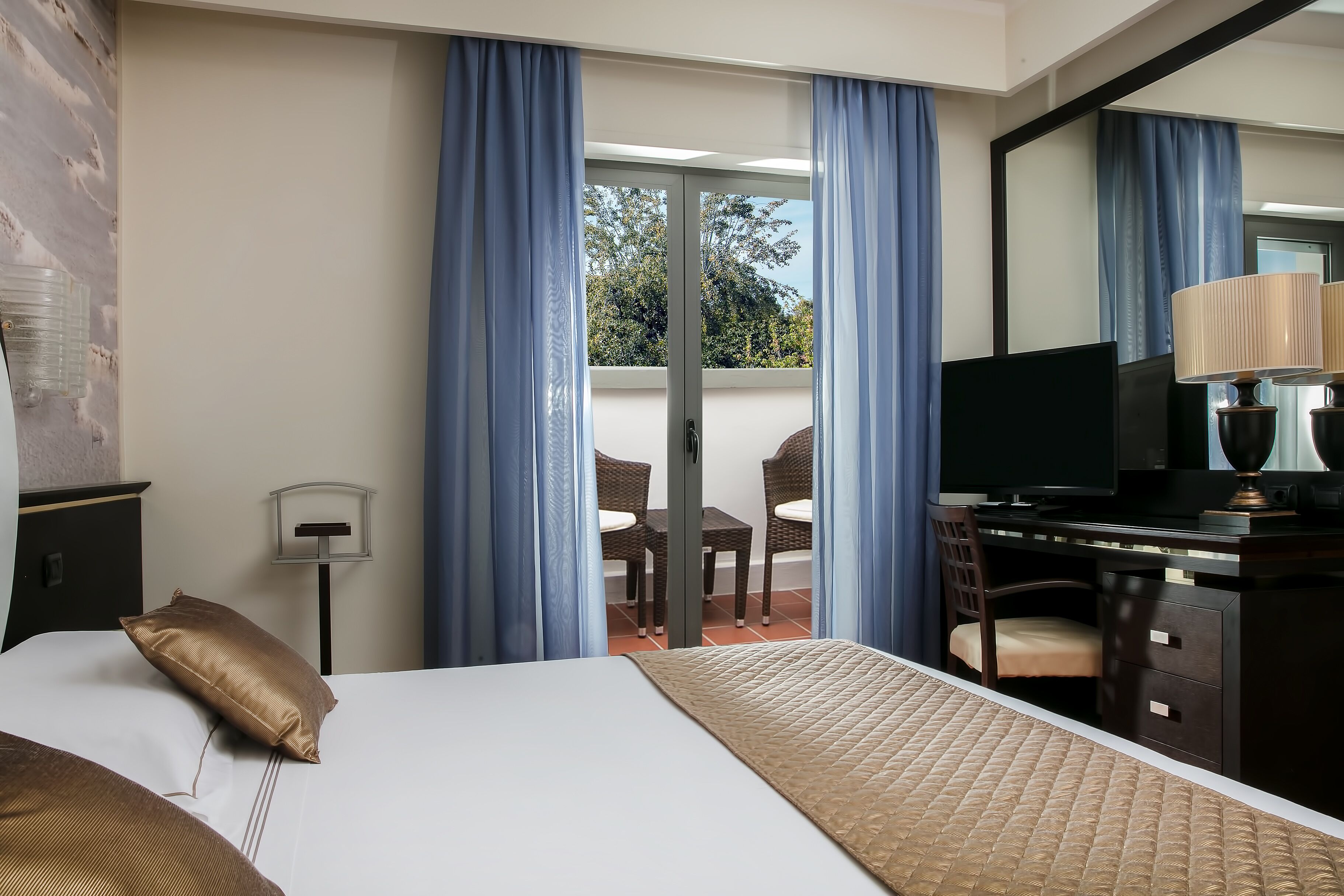 Habitación de hotel con cama, escritorio con televisor, silla y balcón con cortinas azules.