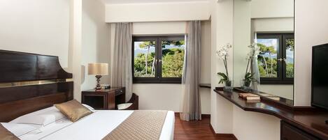 A modern hotel room with a large bed, a desk with a lamp, a TV, and a window with a view of trees.