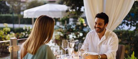 Una pareja cenando en una mesa al aire libre con mantel blanco, rodeada de vegetación y un dosel blanco.