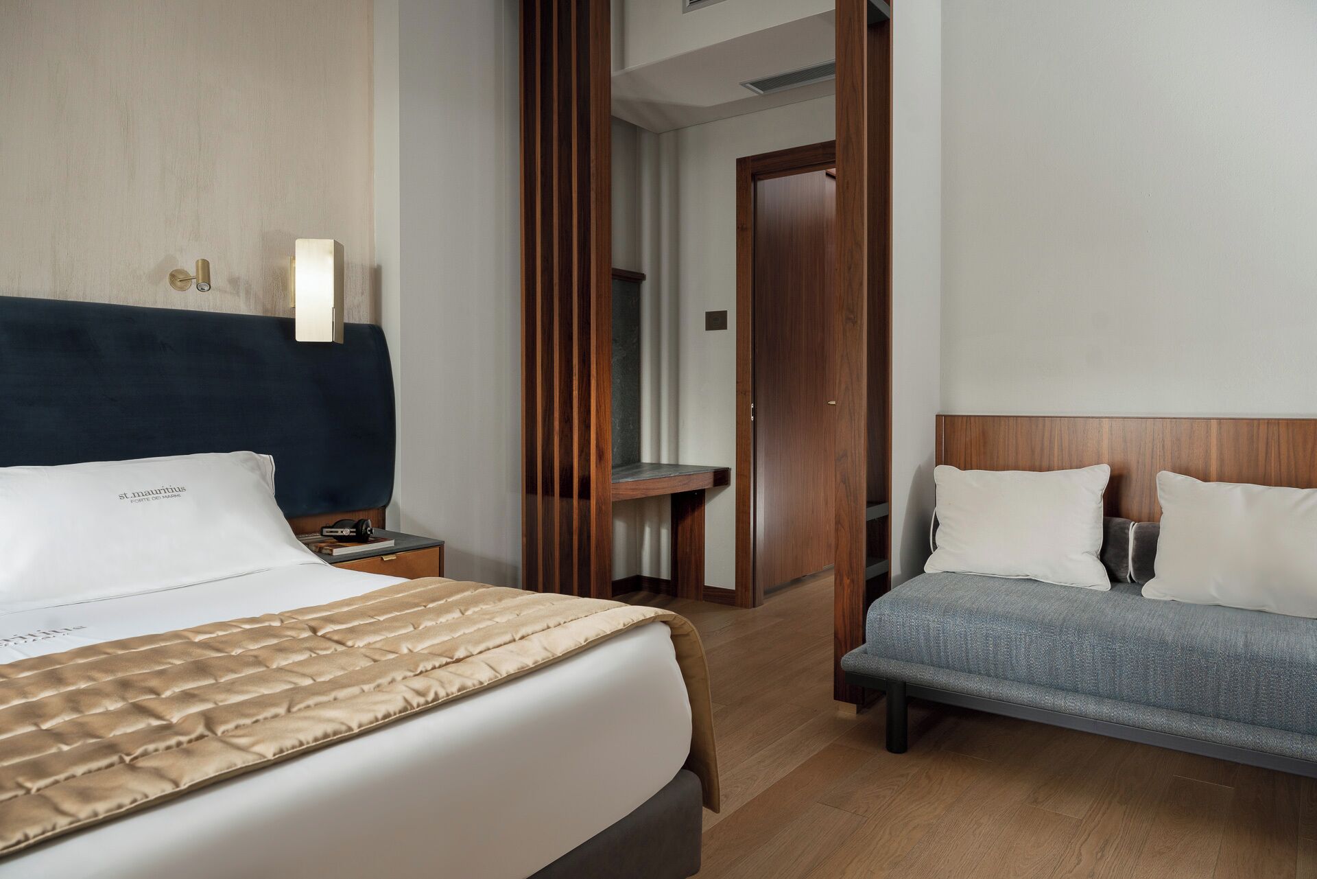 Una habitación de hotel con una cama grande, una cama más pequeña, una mesita de noche, un cabecero de madera, una puerta de madera y un banco.