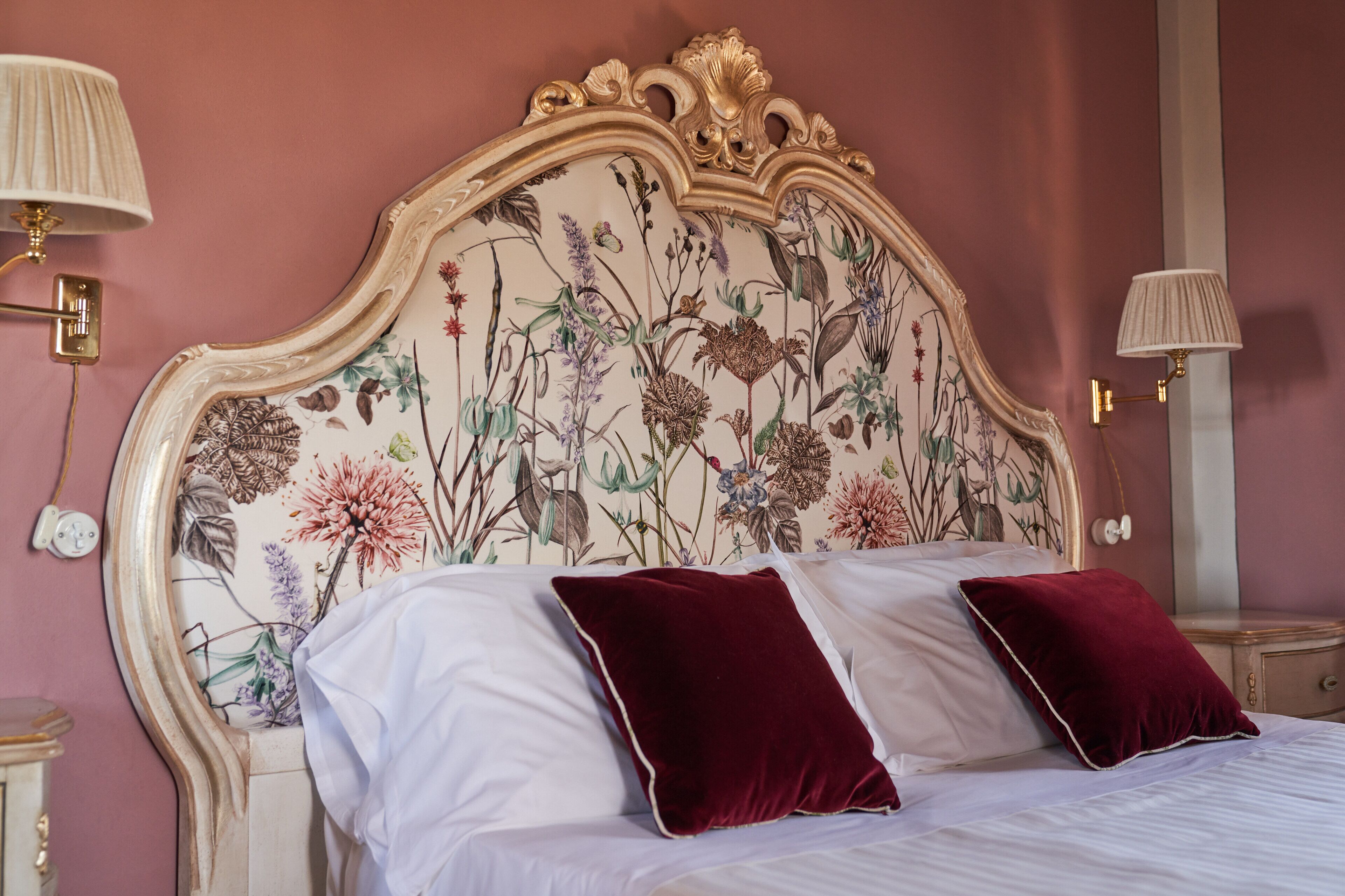 A bed with white linens and two red pillows, a floral headboard, and two bedside lamps.