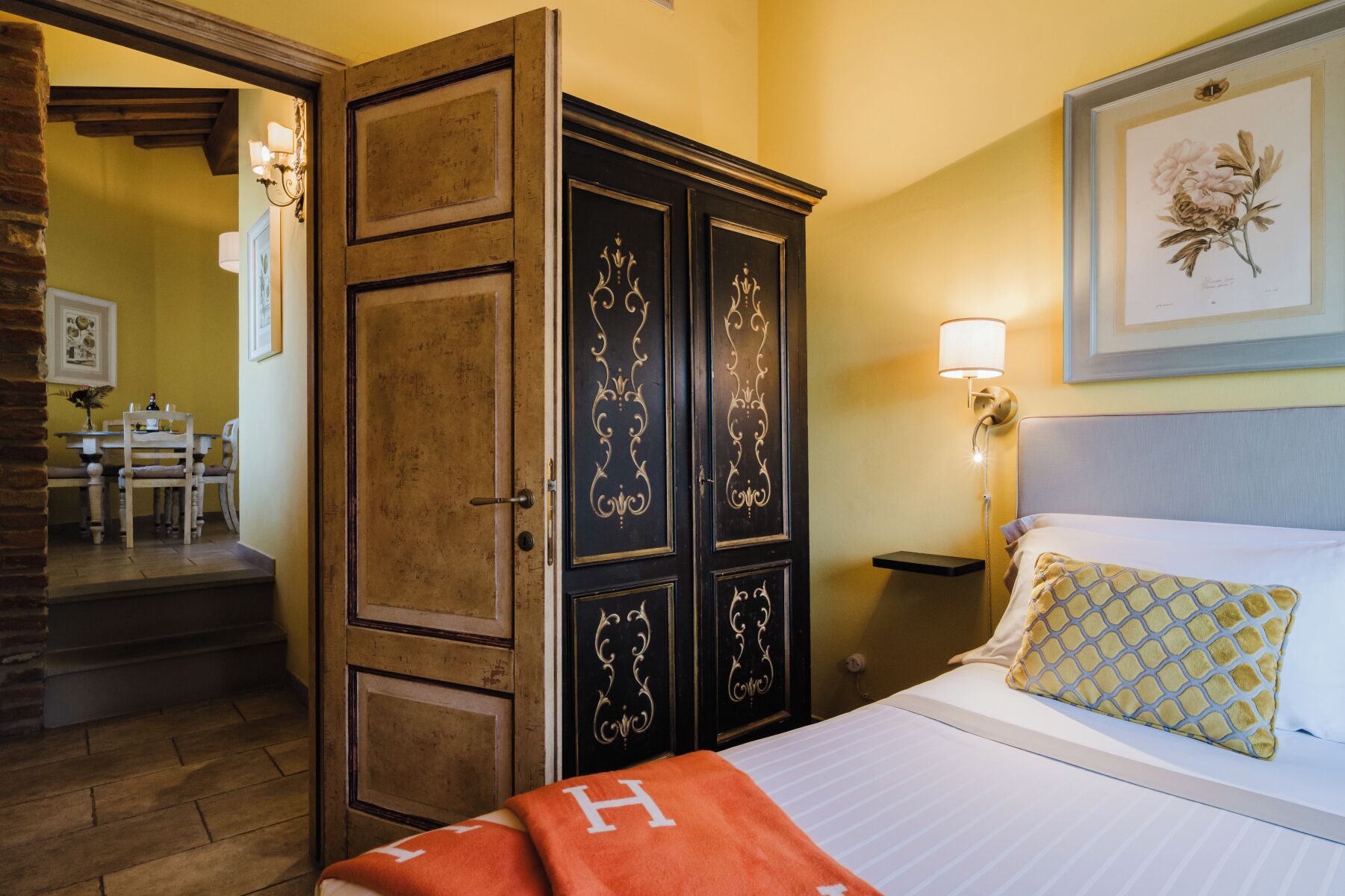 Una camera d'albergo con un letto, un comodino, una lampada, un quadro floreale incorniciato e un armadio in legno.