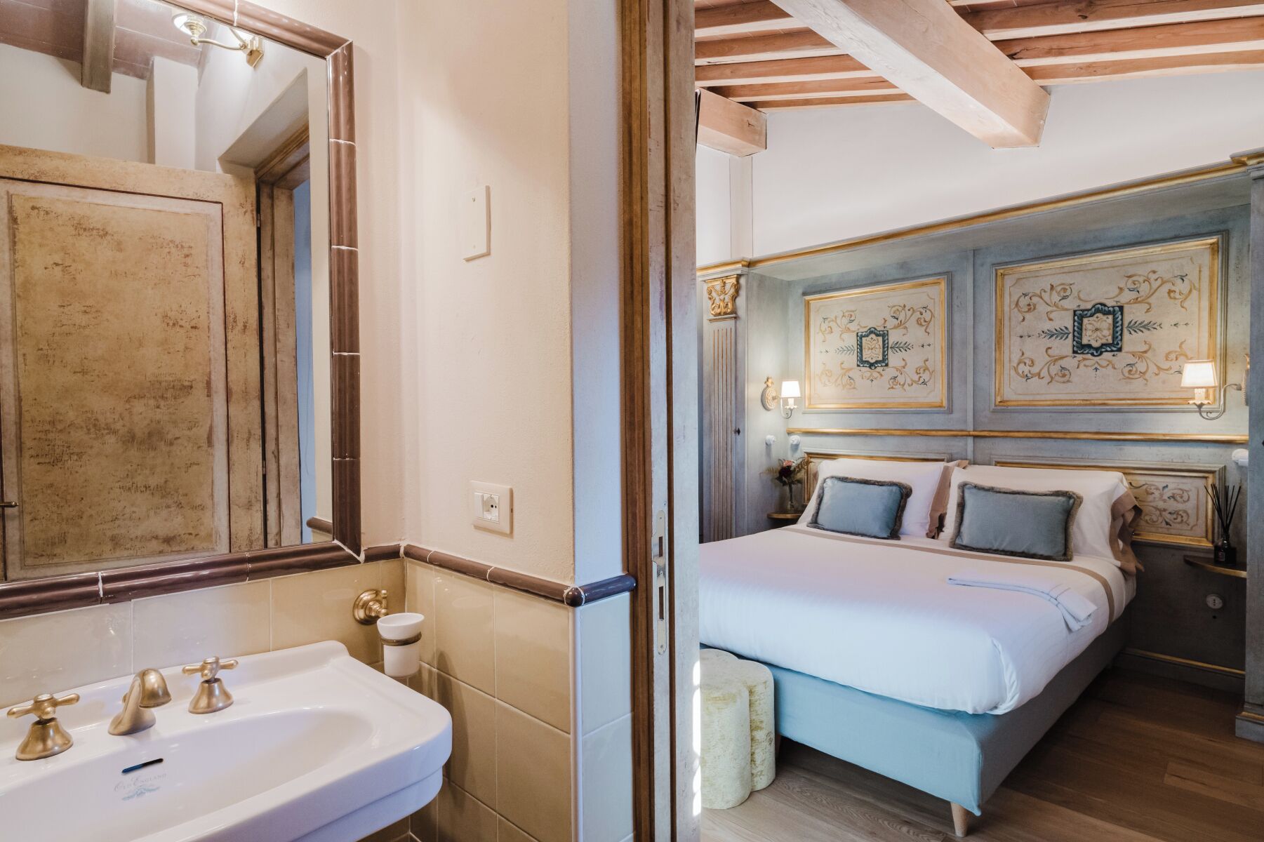 A bathroom with a sink, mirror, and toilet. The room has wooden beams on the ceiling and a bed with a headboard and pillows.